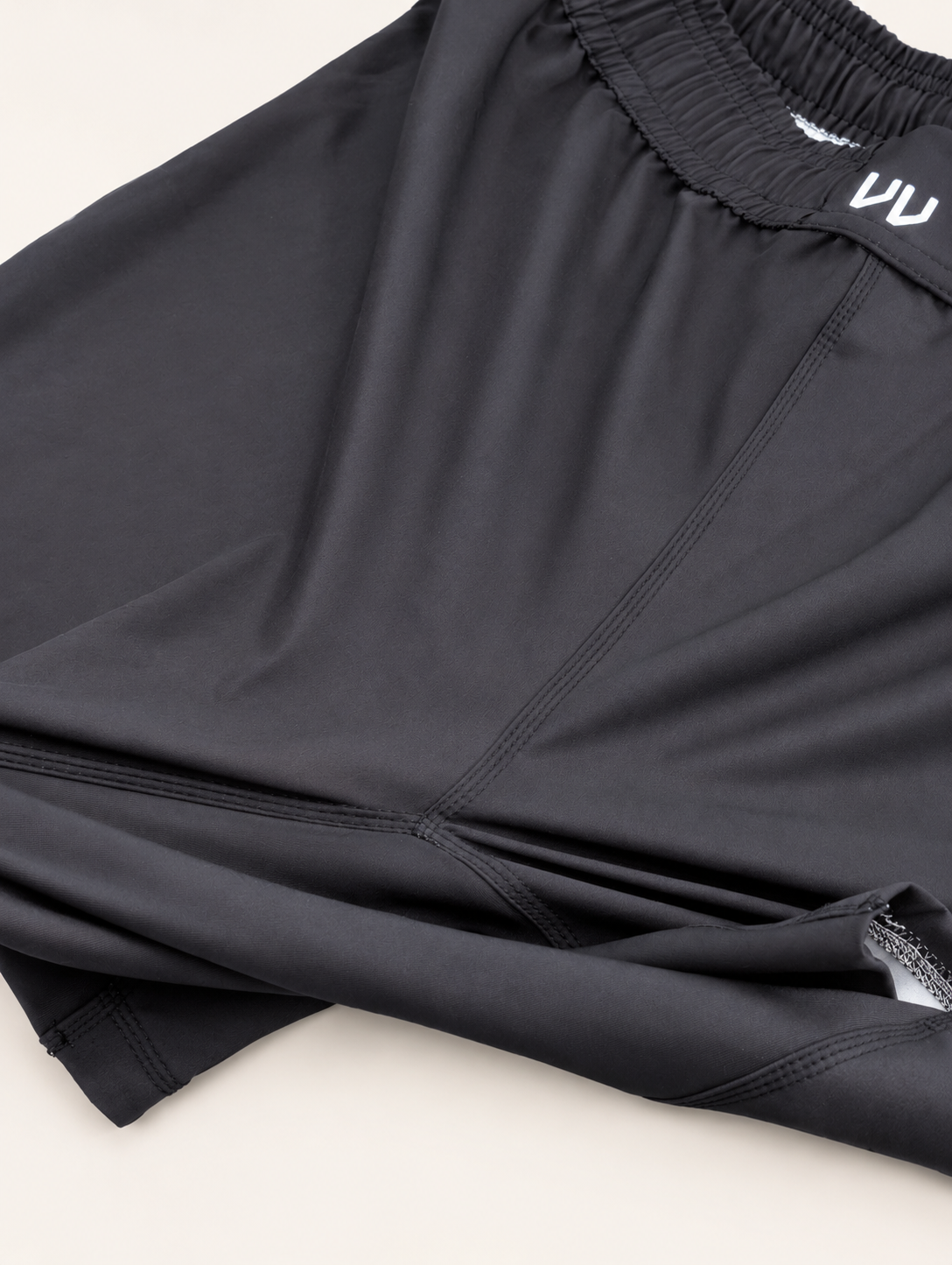 Close-up of a black athletic top with a gathered elastic neckline and a small white logo on the inside collar, laid flat on a light beige background.